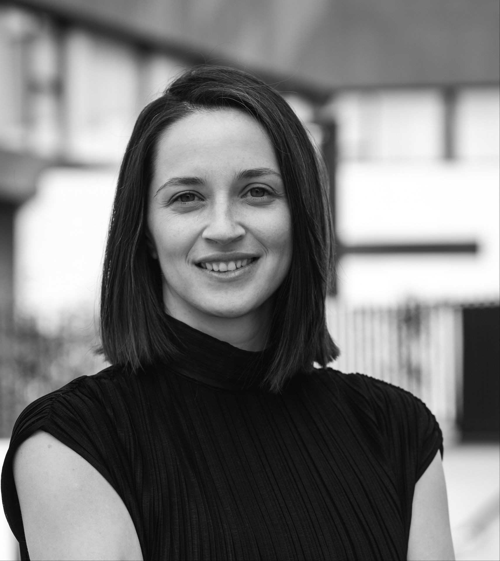 Black and white portrait of a smiling woman with shoulder-length hair, standing in an outdoor setting.